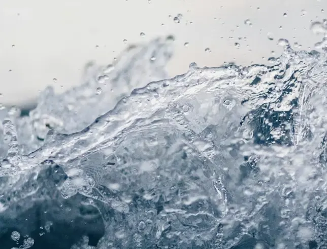 Close-up de água em movimento com salpicos e espuma, sobre um fundo claro e desfocado.